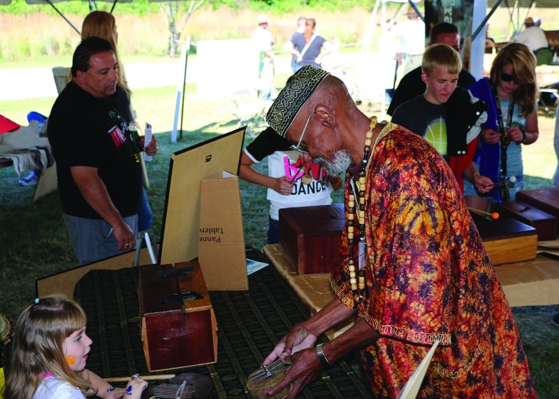 An older man shows a small drum to a young girl while in the background other people look at handmade drums