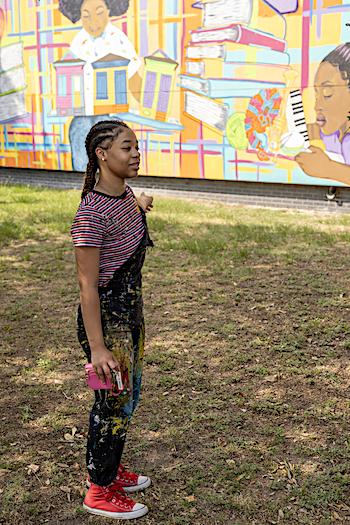 A young woman wearing overalls with paint splatters stands in a grassy area with a large murul in the background
