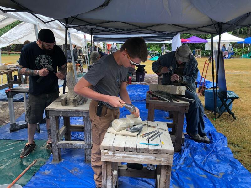 Three men stand at wooden tables under a tent carving wood pieces. 