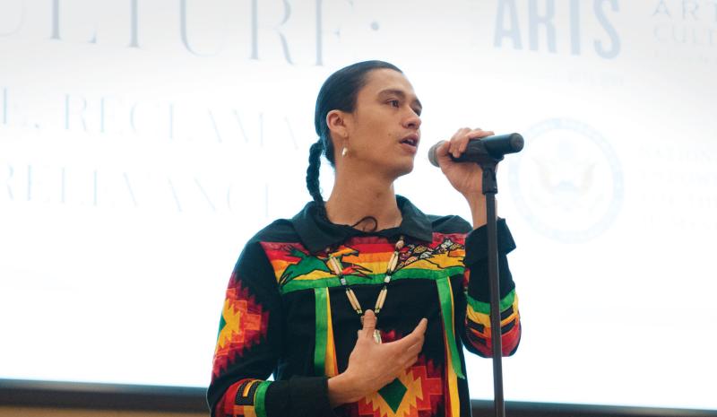 Native man with hair in braid, wearing colorful traditional clothes, at a microphone on stage. 
