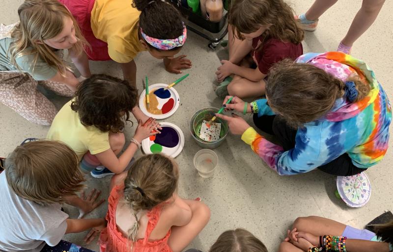 A group of children gather with a facilitator in a circle with paint and brushes at the center.