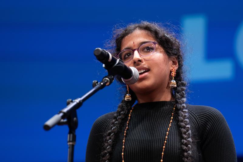 A young woman with glasses and two long braids speaks into a microphone