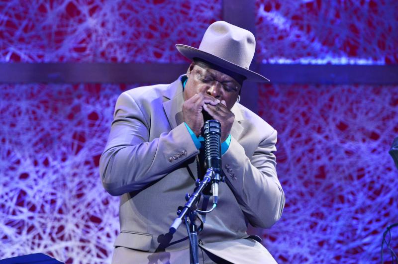 A close-up of a man wearing a light brown suit and hat, playing a harmonica that is cupped in both his hands. He is sitting on a stage behind a microphone. His eyes are closed as if he is 100% focused on the music.