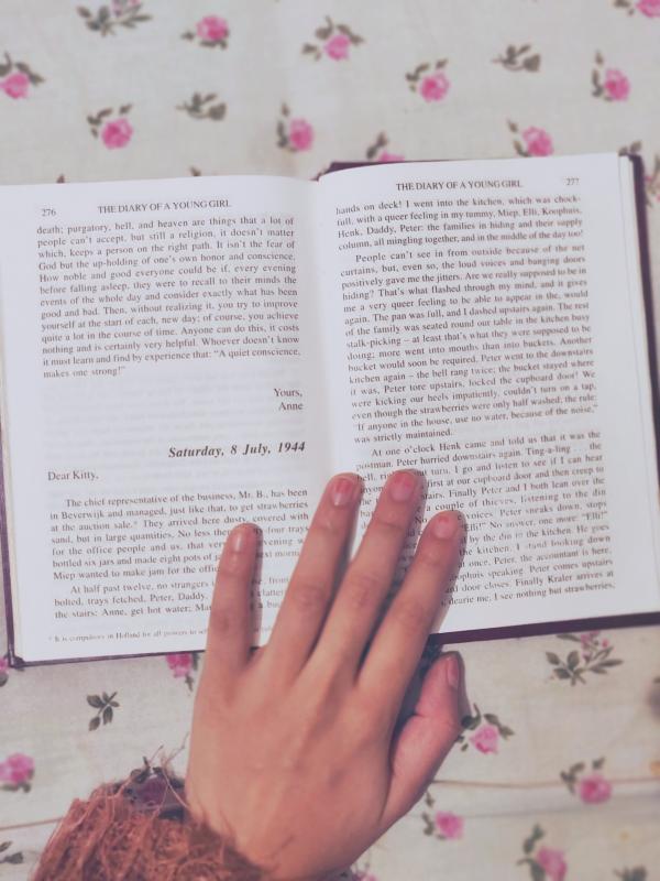 White hand touching a chapter within Anne Frank's book: The Diary of a Young Girl on top of a table that has white and floral decor.