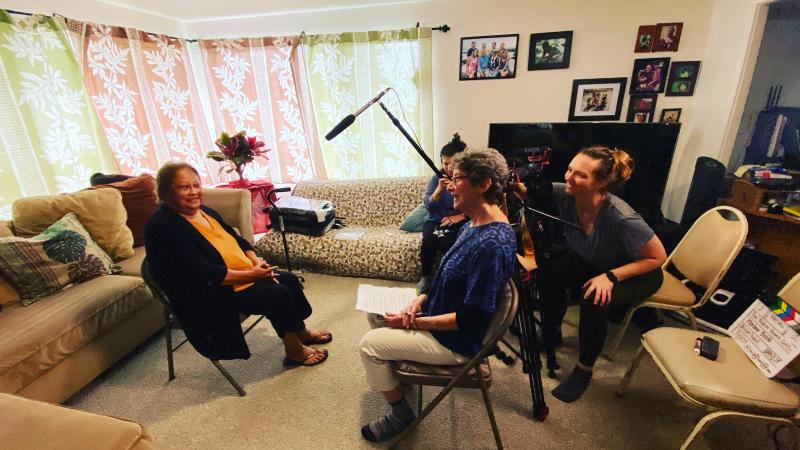 Three women sitting in chairs filming an elderly Hawaiian woman. 