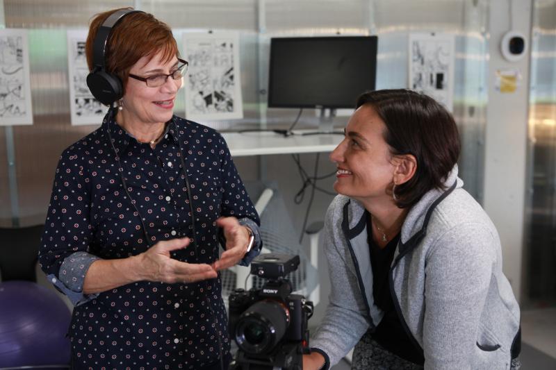 Left: Woman with short red hair and a blue button down shirt holding a camera and wearing headphones. Right: Woman with earl length brown hair wearing a grey goodie with headphones around her neck.