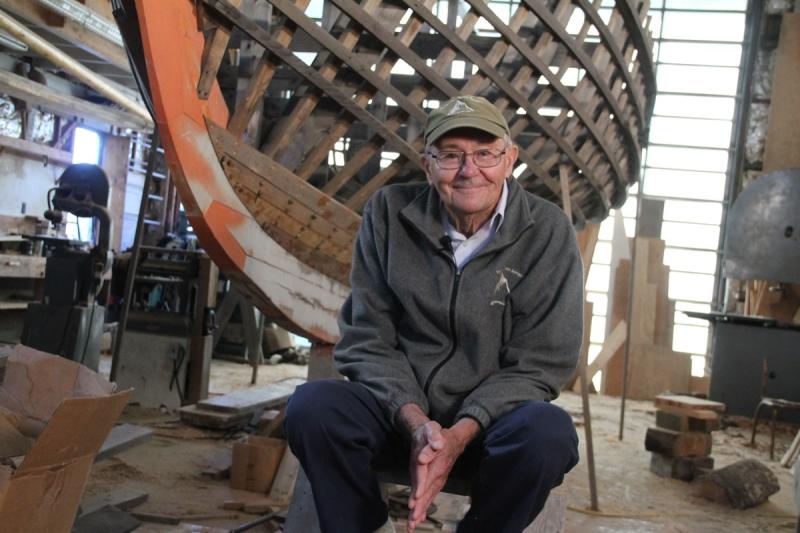 Man sits on a stool in front of the frame of a wooden boat during construction