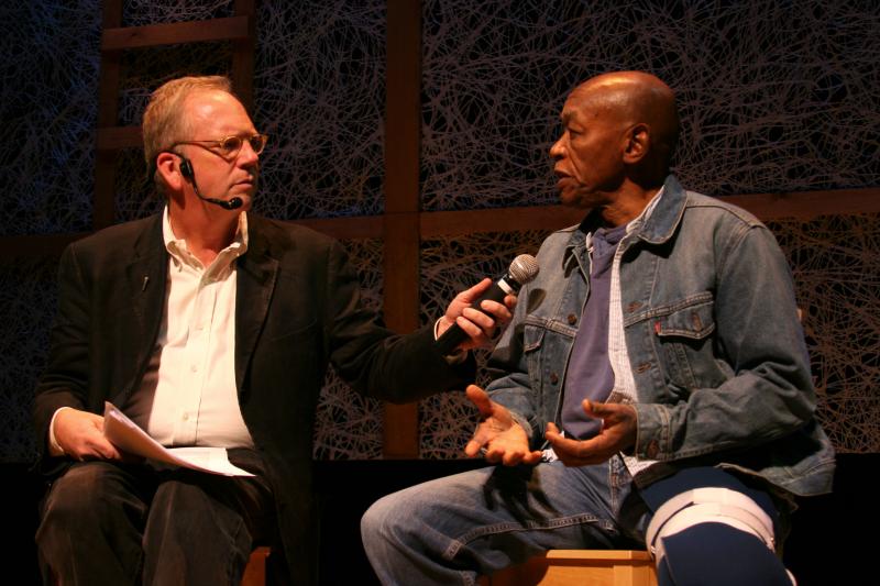 Two men sitting on a stage talking with one another. The man on the left is holding a microphone up for the man on the right