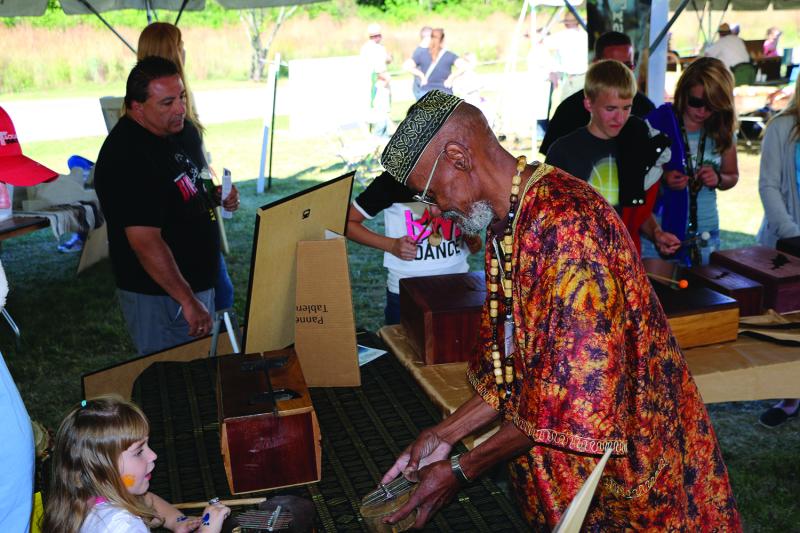 An older Black man in a colorful traditional African outfit shows a drum to a young white child