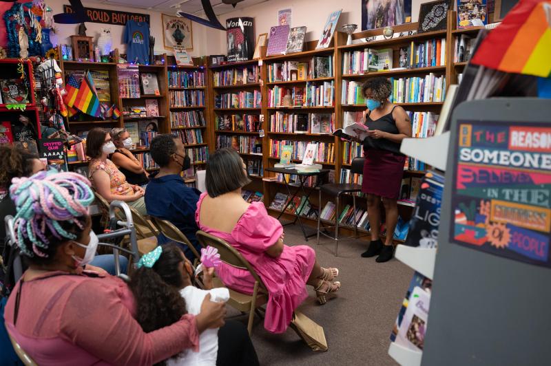 Woman in a mask stands in front of a wooden bookshelf filled with books and reads a book to an audience. Woman in a mask stands in front of a wooden bookshelf filled with books and reads a book to an audience.