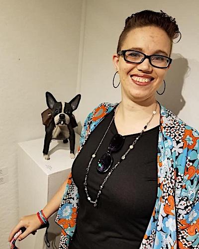 Shawna N.M. Barnes is a white woman in her late thirties with short brown hair, glasses, and hoop earrings. She is smiling and wearing a black shirt with a blue floral cardigan, and has a necklace and sunglasses around her neck. She is holding onto a cane/walker and is standing in front of a sculpture of a dog against a white background.