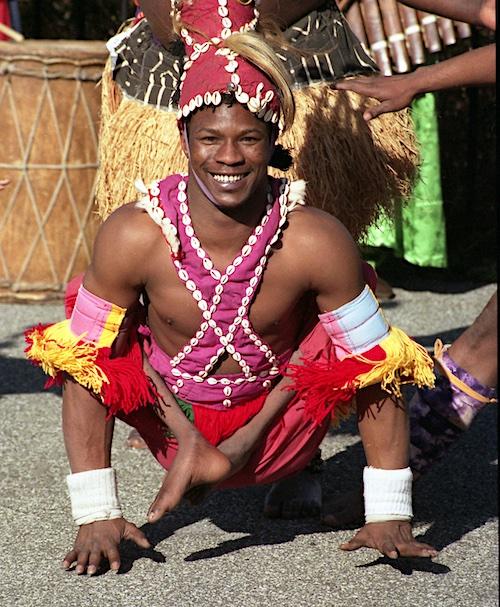 Sidiki Conde is a black man in his thirties. In this photo he is smiling straight at the camera, balancing on his hands holding himself up with his legs crossed at his sides. He is wearing red, yellow, pink and brown striped frilly cloth on his right arm where his elbow is, and one on his left but it is a light blue. He is wearing a hat-like ornament with shells on his head which also crisscrosses across his bare chest. He wears red shorts, and two wrist bands on each wrist.
