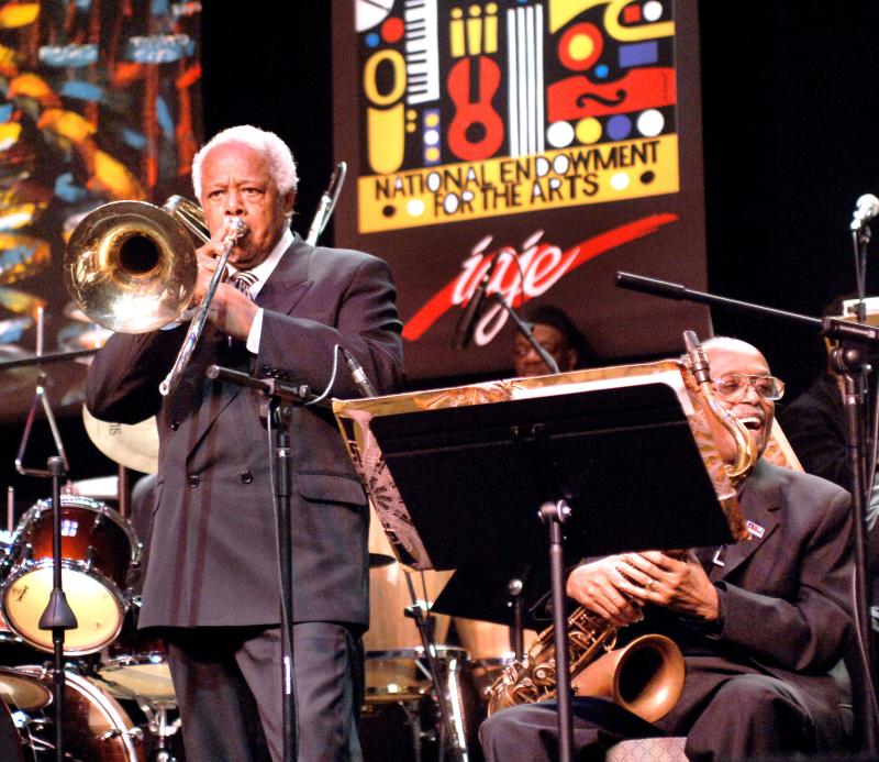 A man stands playing the trombone, next to him is a man seated, holding a saxophone