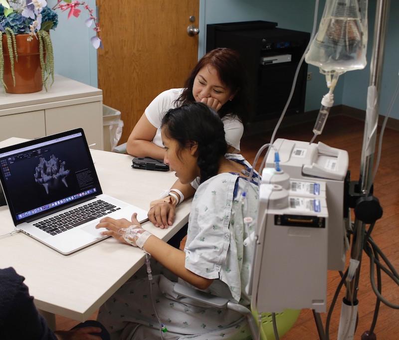 a young patient wearing a hospital gown interacts with a computer screen while an adult looks on
