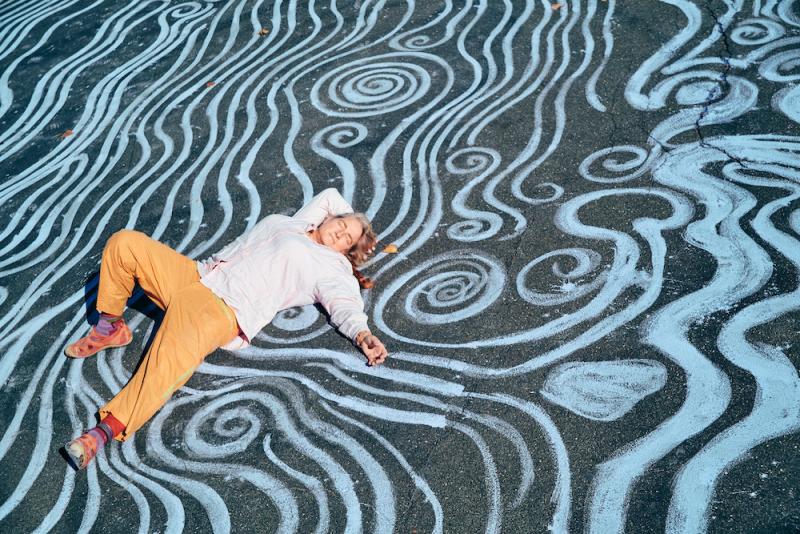 Stacy Levy, who is a white woman, lays on a strip of pavement colored to look like rivulets of water