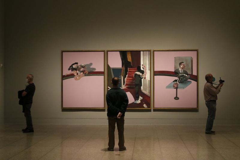 Masked museum visitors look at three large paintings by Francis Bacon