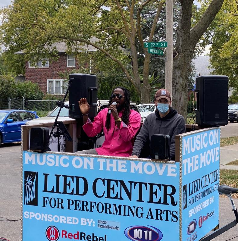 A musician and a man in a mask perform in a box attached to a bicycle outside