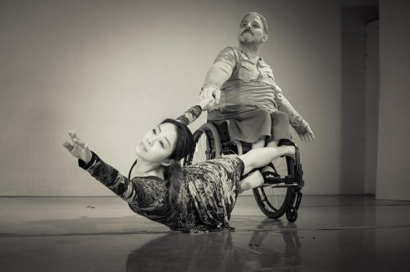 Black and white photo: Dancer on left in a floral dress reaching out into the abyss, with a dancer (in a wheelchair) on the right who is wearing a cap, tshirt, and pants. Black and white photo: Dancer on left in a floral dress reaching out into the abyss, with a dancer (in a wheelchair) on the right who is wearing a cap, tshirt, and pants.