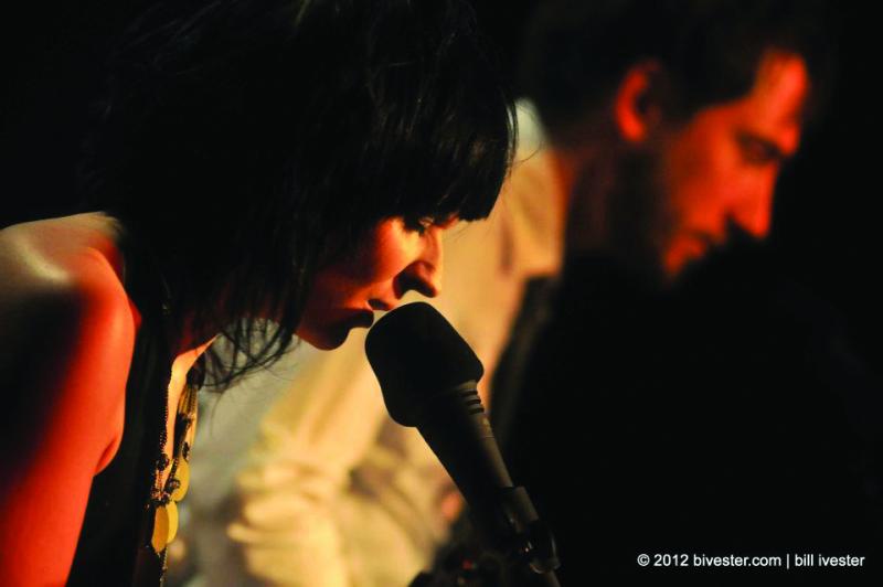 Woman with short black hair singing into a mic with a man out of focus in the background.