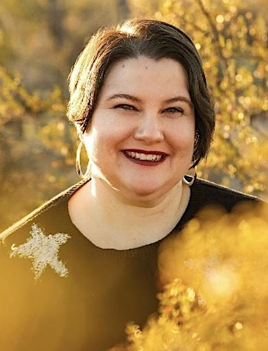 Light-skinned Mestize with dark hair, silver hoop earrings, burgundy lipstick and a black sweater with a white star sits in their scooter smiling surrounded by golden creosote bushes.