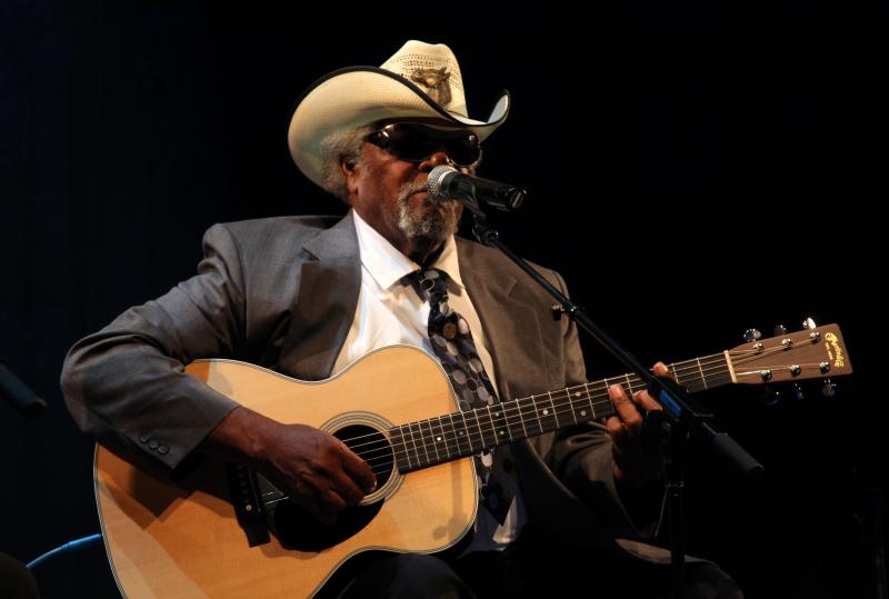 A man in sunglasses and a cowboy hat plays guitar and sings