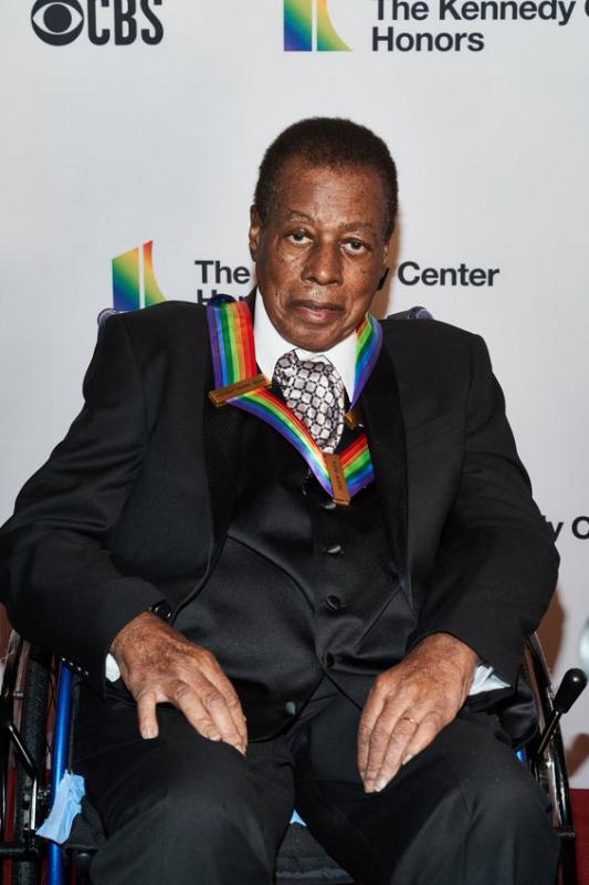 Black man in gray suit in wheelchair with medal with rainbow colored bow around his neck.