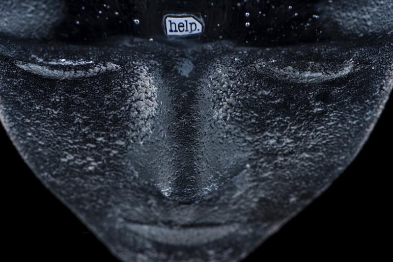 detail from a mask which is painted black and has a scrap with the word help embedded in the forehead of the mask