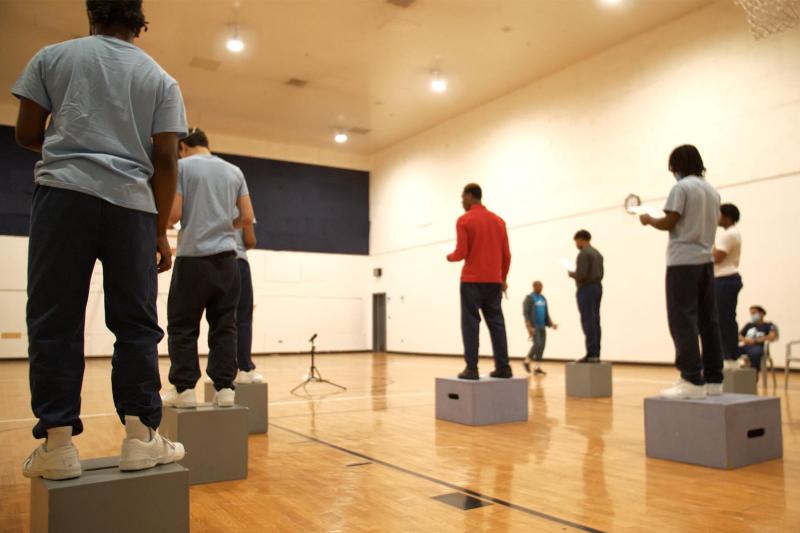 In a gym facing away from the camera: Seven young men (wearing sweatpants and shirts), stand on grey boxes. In a gym facing away from the camera: Seven young men (wearing sweatpants and shirts), stand on grey boxes.