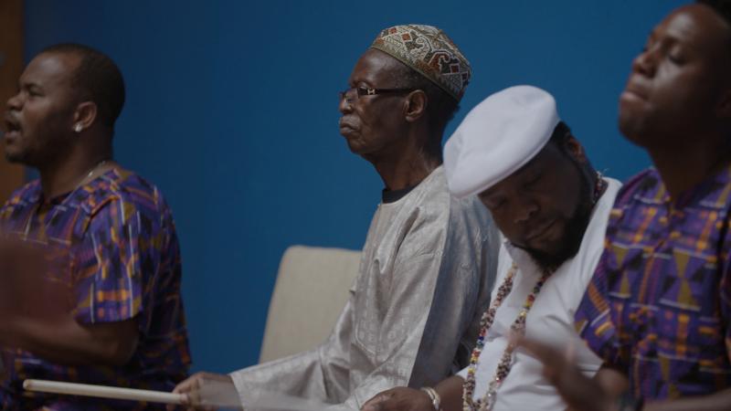 Zak Diouf_photo credit Hypothetical Films Four men sit playing drums with a blue wall in the background