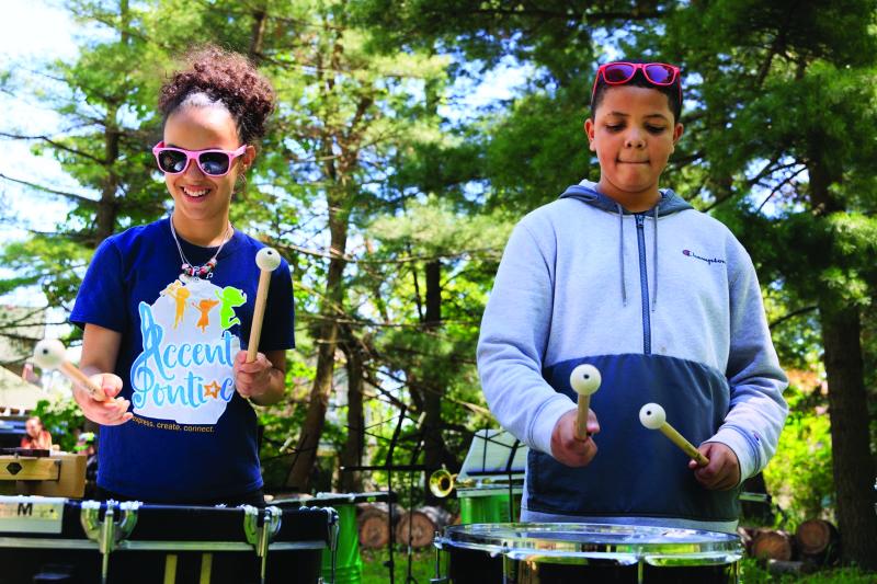 A girl in sunglasses and a boy in a sweatshirt playing drums on a stage outdoors.