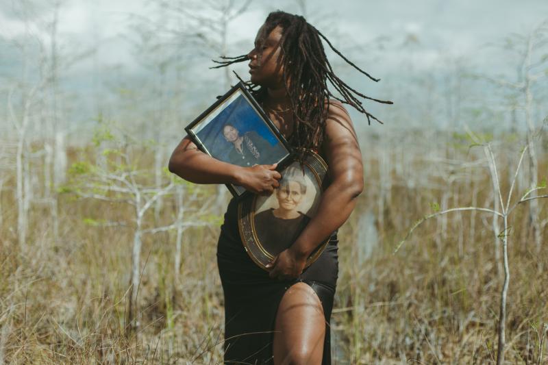 A Black woman holds two photos in vintage frames in the midst of a swamp landscape