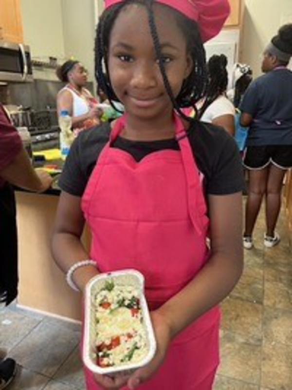 A young Black girl (in a red chef hat and apron) holds a pan of food.