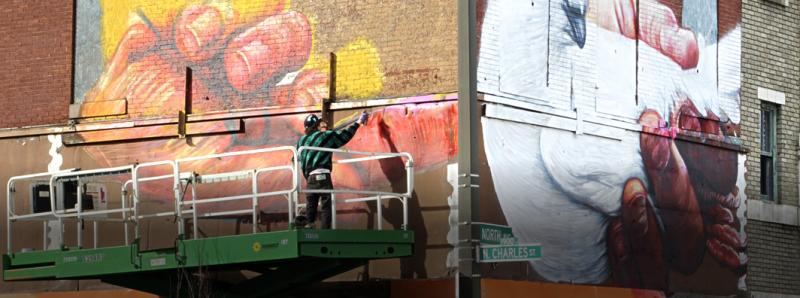 Artist on a platform working on a huge mural of a handion the side of a downtown brick building