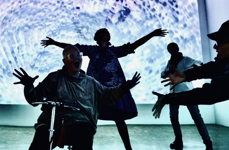 Petra, with her mouth wide open, her collaborator and wife Stephanie Heit next to her, and a number of community participants all dancing in the light of Becoming Fossil, a community dance video about climate emergencies and resiliences. The screen shows a color-shifted close-up of a fossil coral.