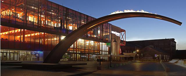 Night shot of a large steel arch with flames in front of a fully lit up multi-story glass buildingfront