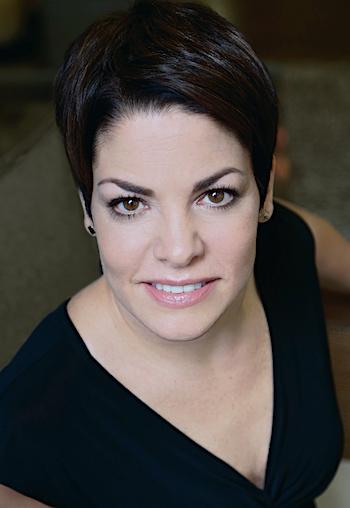 A petite, cis, white woman with short, dark brown hair and brown eyes. She sits in an overstuffed green chair and wears a black V-neck dress with short sleeves and small black square earrings. She smiles directly into the camera.