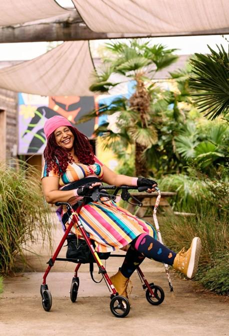 African American woman with auburn locs in a rainbow dress and cool adaptive wear sitting in a red rollator and holding a cane covered in stickers