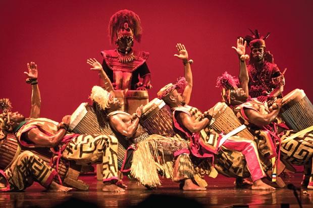 African drummers onstage in traditional costume leaning back in a dance pose with a tall man behind against a red background