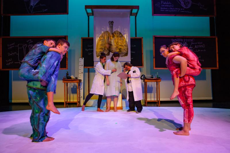 Seven actors are on stage with the settings displaying medical objects and a photo of a heart center stage.