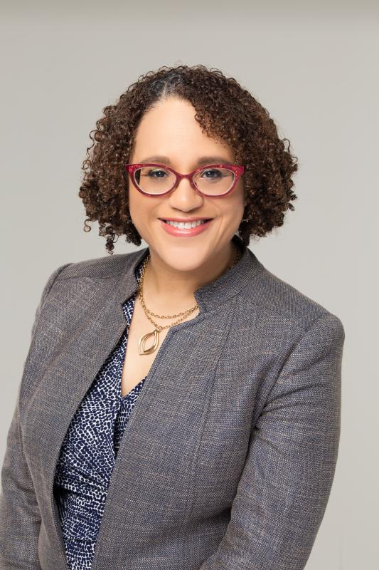 Black woman smiling, wearing red eyeglasses and a grey blazer.