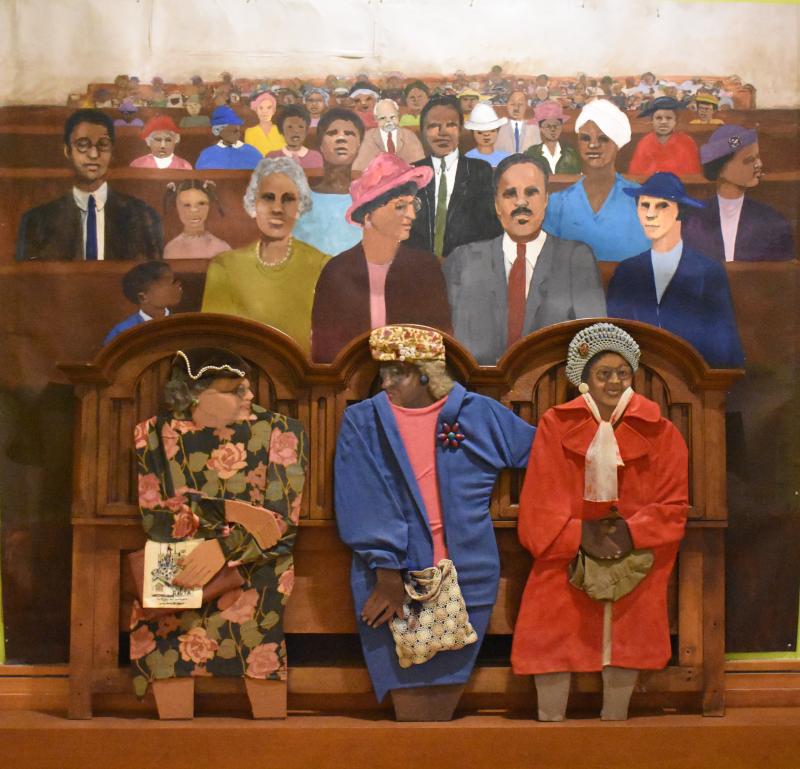 A mixed media work of wood, paint, and cloth of three women in front of a painted backdrop of a group of Black churchgoers.