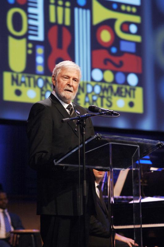 A man with white hair and beard stands at a lectern with a graphic behind him with colorful musical instruments.