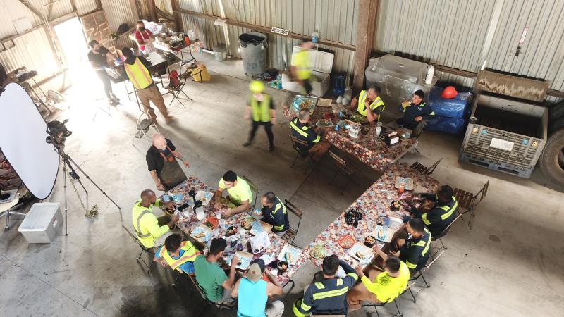 An overhead shot of many people seated around three tables arranged in a "U". They are wearing construction clothes and eating. A cooking station is set up in the upper left corner, and in the bottom left is a camera on a tripod and light reflector/diffuser.
