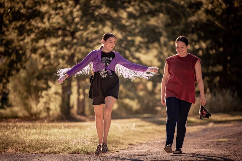 Native teen girl (left) wearing a purple jacket and black shorts and Native woman (right) wearing a red shirt and black pants, walking through a grassy area with trees surrounding them.