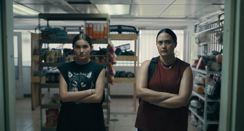 Native teen girl (left) wearing a graphic t-shirt and Native woman (right) wearing a red shirt: are inside of a store with their arms folded in front of them