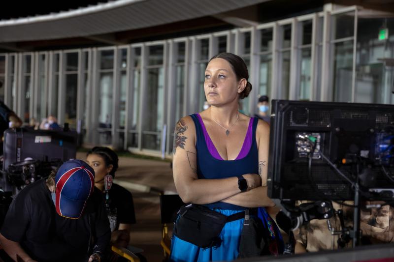 Native woman (center) stands in front of a camera monitor and looks to her right. A woman is seated behind and looking to her right. A man wearing a red, white, and blue hat and is sitting and looking down.