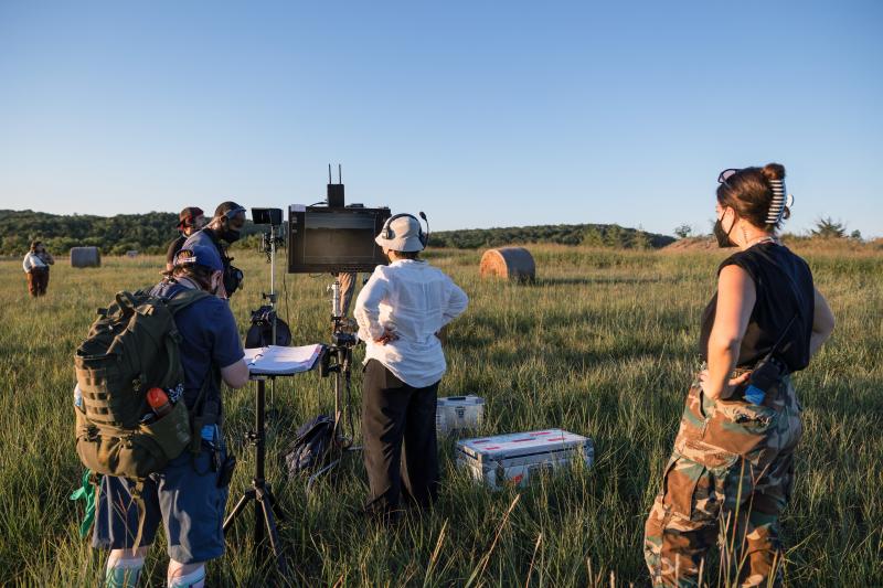 A film crew are standing in a field, facing a camera monitor as they shoot a film scene.