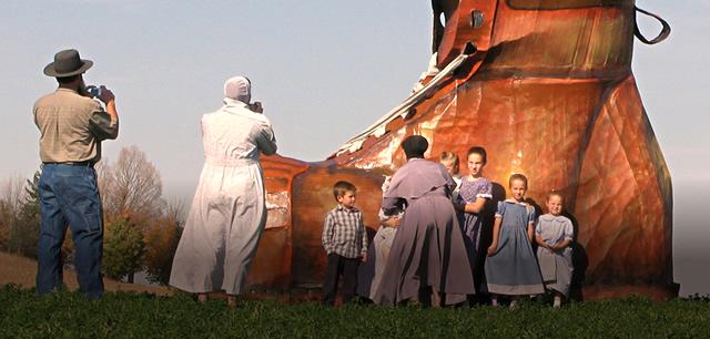 Amish parents and children looking at a giant wooden sculpture of a work boot 