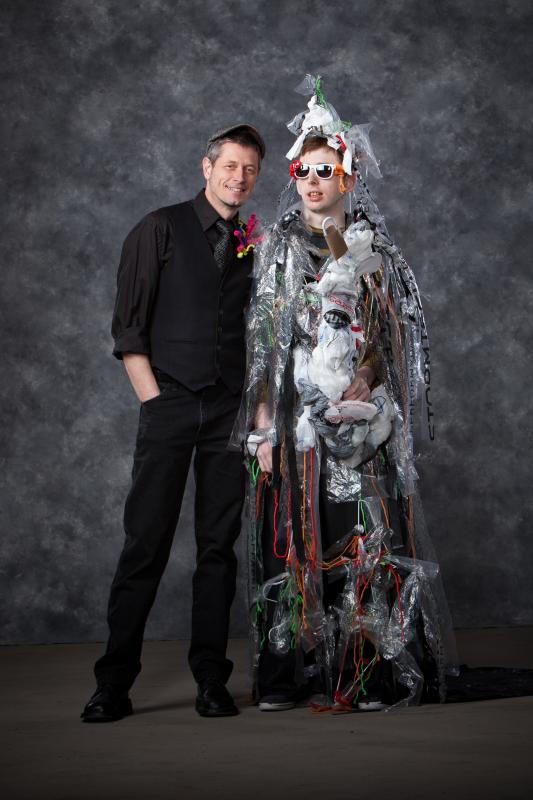 Man (left) wearing a black suit and grey hat on his head. Man (right) wearing sunglasses and an eccentric garbage bag design with various multi-colored material attached to it.