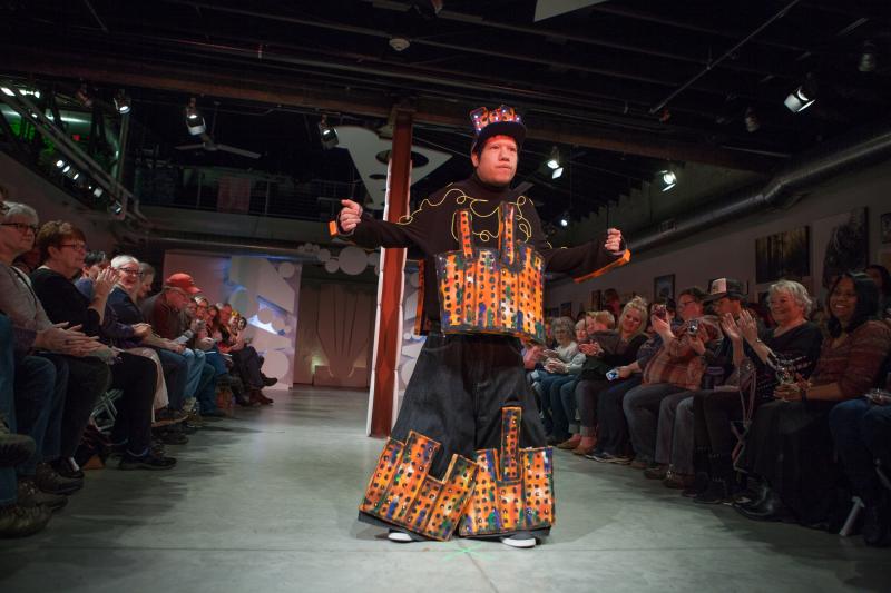 Man on a runway wearing a multi-colored outfit that has images of buildings on it. Audience members are watching on both sides of the runway.